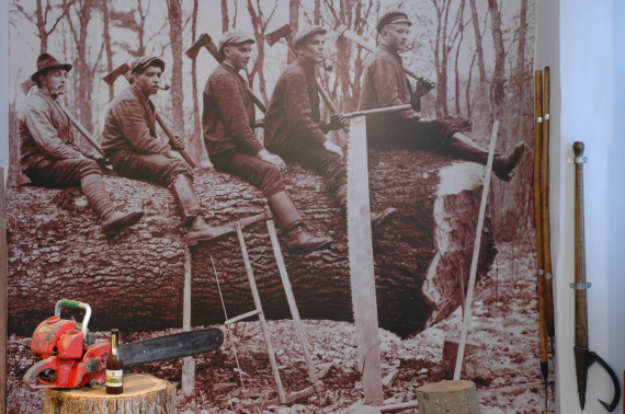 Inszenierung in Raum 101 (Holzernte): schwarz-weißes Großfoto im Hintergrund, das fünf junge Männer zeigt, die jeweils eine Axt über der Schulter tragen. Sie sitzen hintereinander rittlings auf einem gefällten Baum. Davor sind verschiedene Werkzeuge zum Holzfällen aufgebaut, unter anderem eine Zugsäge und eine Motorsäge.