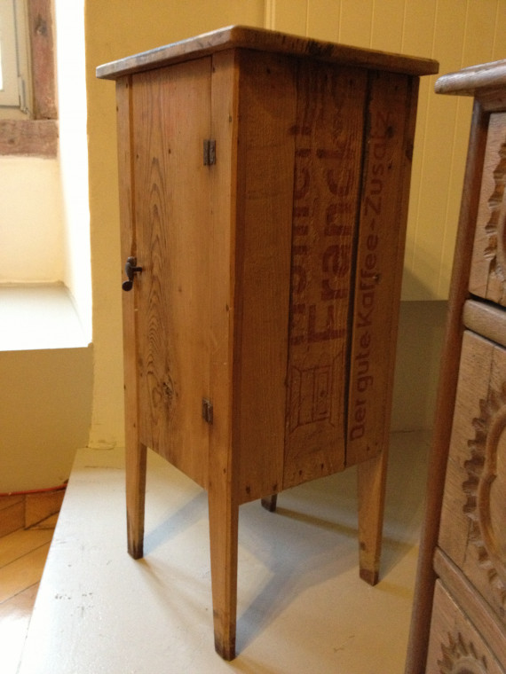 Ein Nachttischschränkchen aus Holz, das ursprünglich eine Kaffeekiste war. Die Holzkiste mit roter Werbeaufschrift wurde auf die Seite gedreht und mit vier einfachen flachen Holzbeinen versehen.