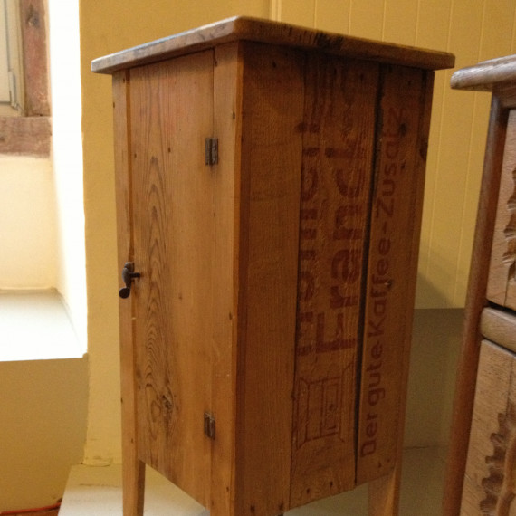 Eine alte Kaffee-Kiste aus Holz mit der Aufschrift &quot;Franck. Der gute Kaffee-Zusatz&quot;. Die Kiste wurde auf die kurze Seite gestellt und mit schlichten, geraden Holzbeinen versehen. Jetzt ist sie ein Nachttischschränkchen.